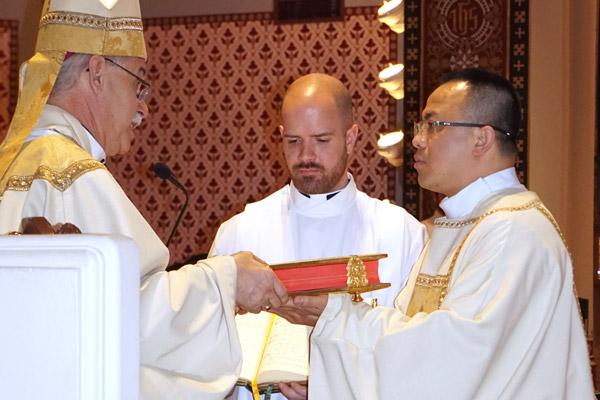 Bishop Taylor presents Deacon Tuyen Do with the Book of Gospels as seminarian Patrick Friend assists. (Aprille Hanson photo) 