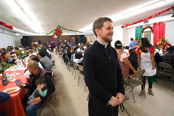 Pastor Father Stephen Hart pauses for a photo during the fiesta at the Ashley County Fairgrounds following the Our Lady of Guadalupe feast day Mass while a Russellville band called Tamborazo Zacatecano de Arkansas performs at the other end of the hall. (Malea Hargett photo)
