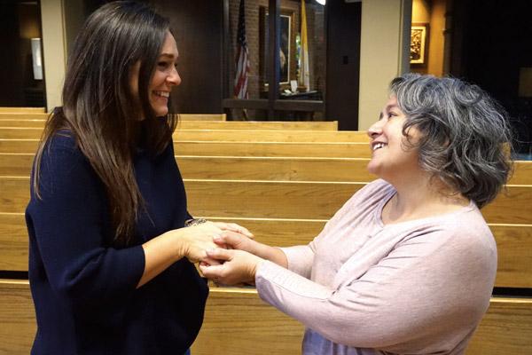 Susie Williams (left) and Stacey Matchett shake hands to demonstrate the typical sign of peace the laity give at Mass. A form of “peace be with you” is also spoken. (Aprille Hanson)