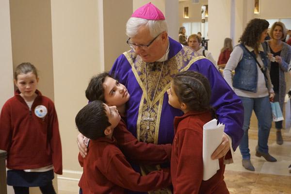 Bishop-elect Francis I. Malone hugs fourth-grade students Addison Cole (left embrace), Sam Cruse and Mary McConnell, as third-grader Maggie McNulty watches. He was prayed over and praised by students at the final school Mass Dec. 20. (Aprille Hanson photo) 