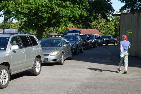 A line of cars wait for their online Farm Stand orders as volunteer Michelle Shellabarger walks to deliver an order to a vehicle. All volunteers wore masks and gloves. (Aprille Hanson photo)