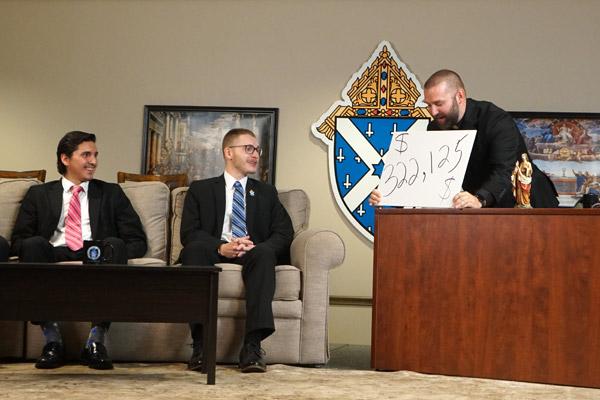 Father Jeff Hebert shows the audience that $322,125 had been raised for seminarian education by the end of the Taste of Faith fundraiser Aug. 7. New seminarians Pedro Alvarez (left) of Little Rock and Joshua Osbourne of Conway look on. (Malea Hargett photo)