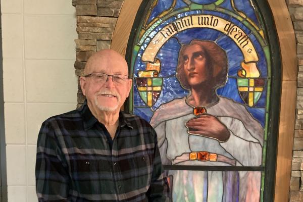 Charlie Ray, 80, stands by a stained glass window at his house. A talented craftsman, Ray built the frame and put in all the stonework, and works on construction projects at All Saints Church in Mount Ida. Courtesy Diane Campbell.